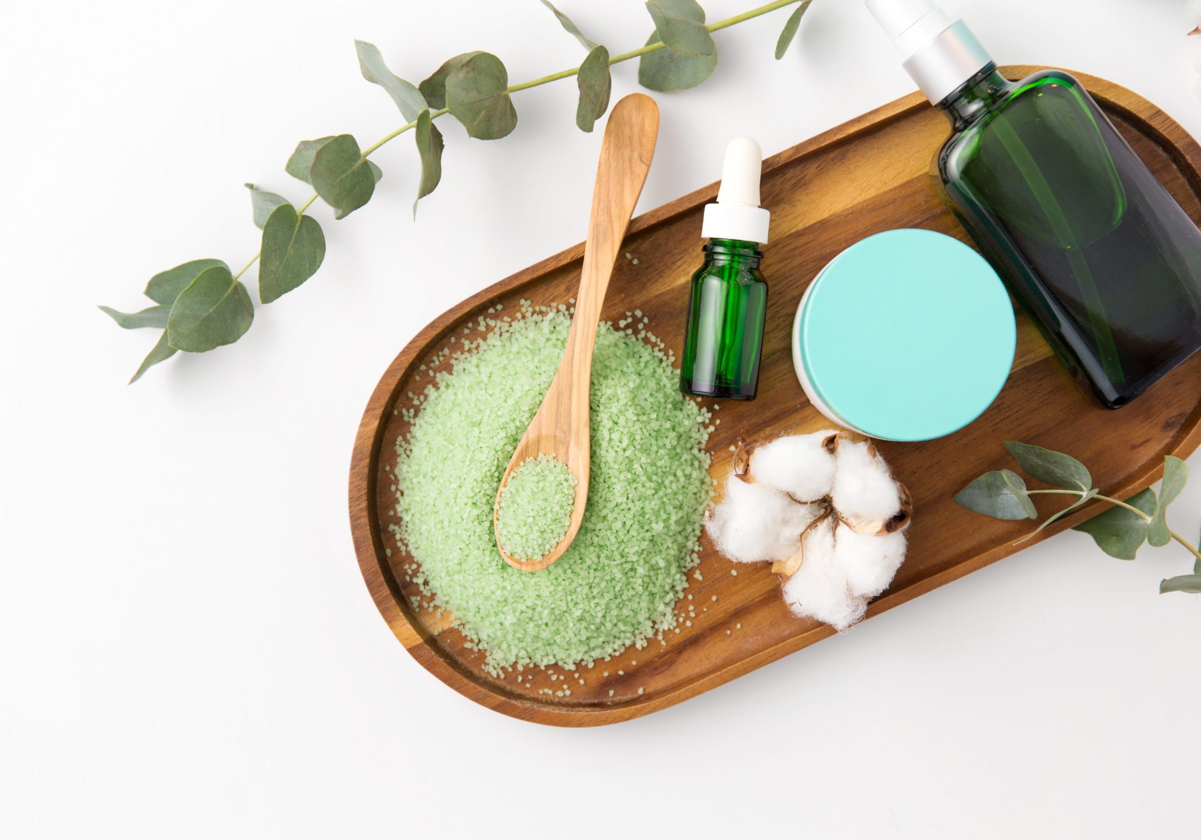 beauty and spa concept - green bath salt, serum with dropper, body oil, moisturizer and eucalyptus cinerea with cotton flowers on wooden tray